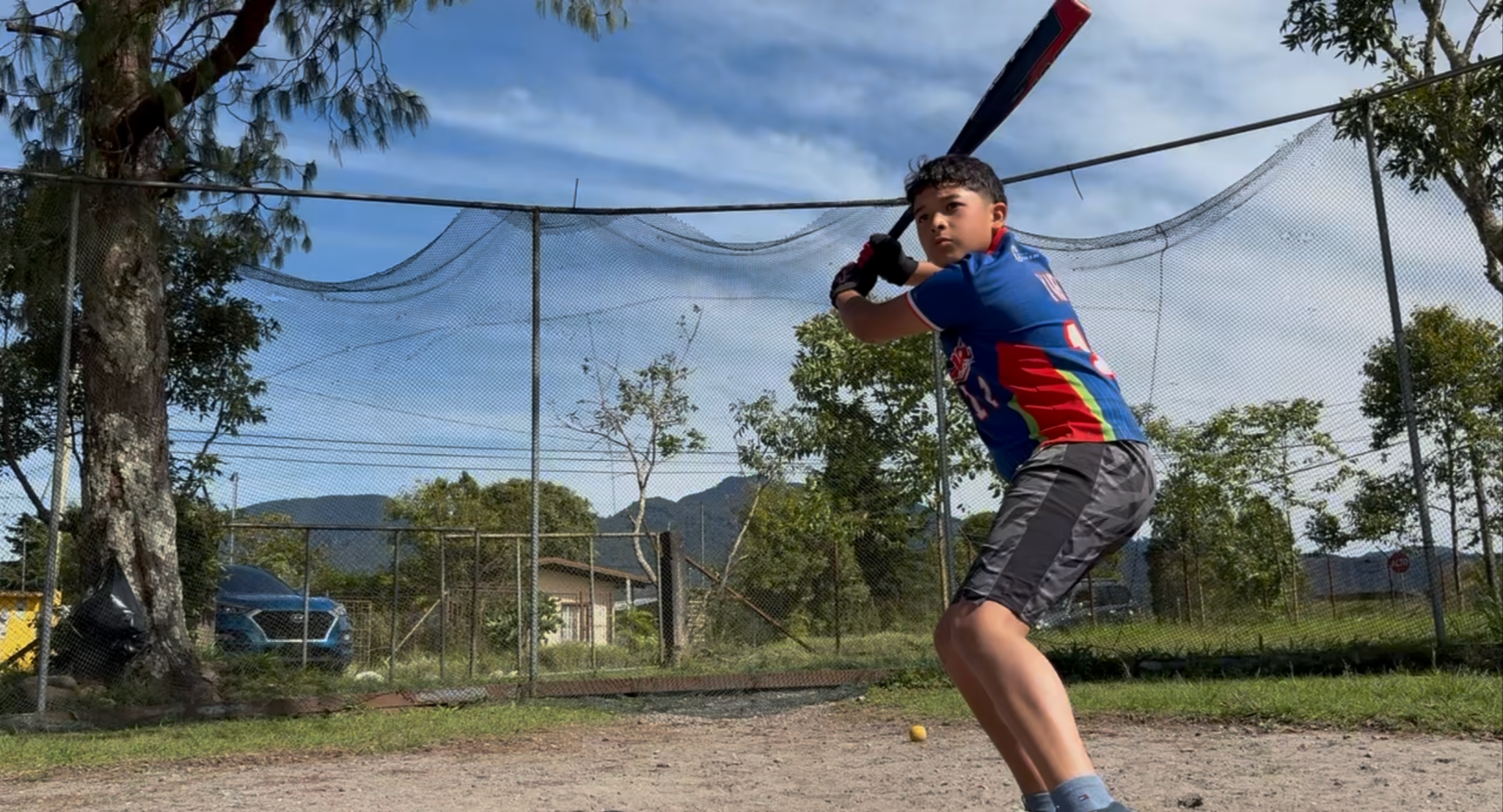 Young boy in a sports jersey and shorts practicing baseball batting outdoors on a dirt field with a chain-link fence and trees in the background.