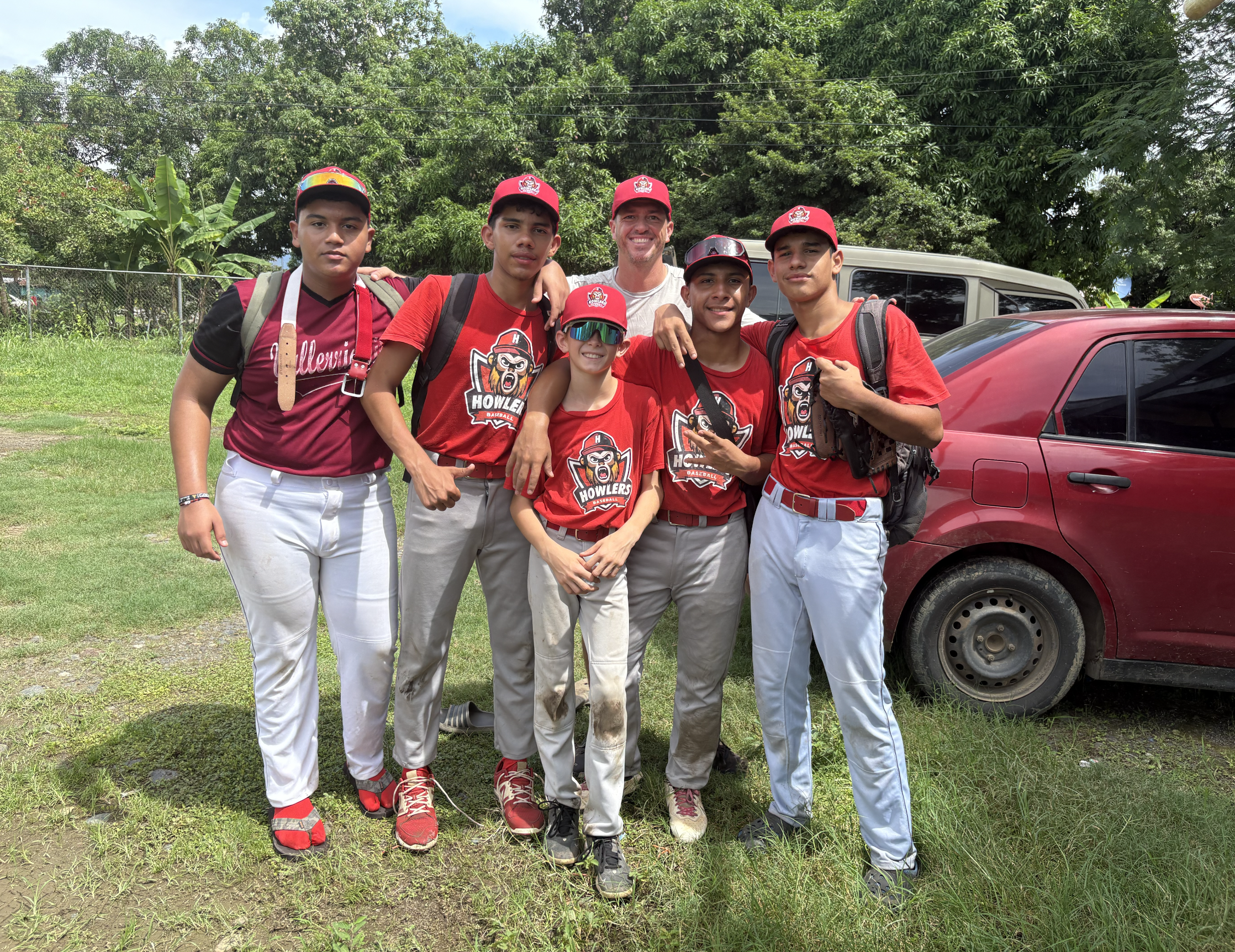 Group of six young people and one adult posing outdoors on grass, wearing red and white sports uniforms with teams logos, some with backpacks and sunglasses, in front of cars and trees.