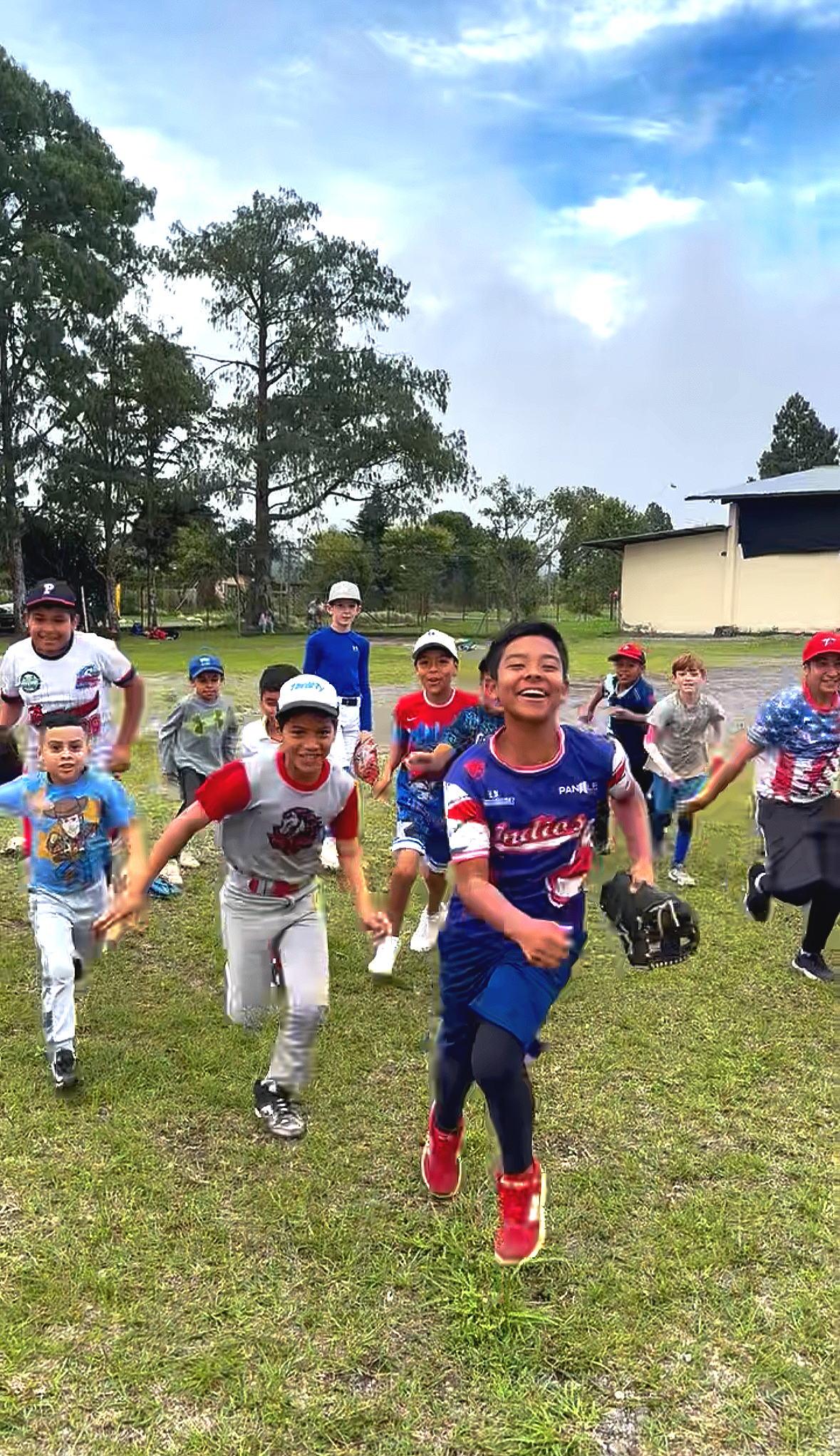 Children playing and running happily outside on a grassy field, some wearing baseball caps and sports jerseys, with trees and a building in the background.