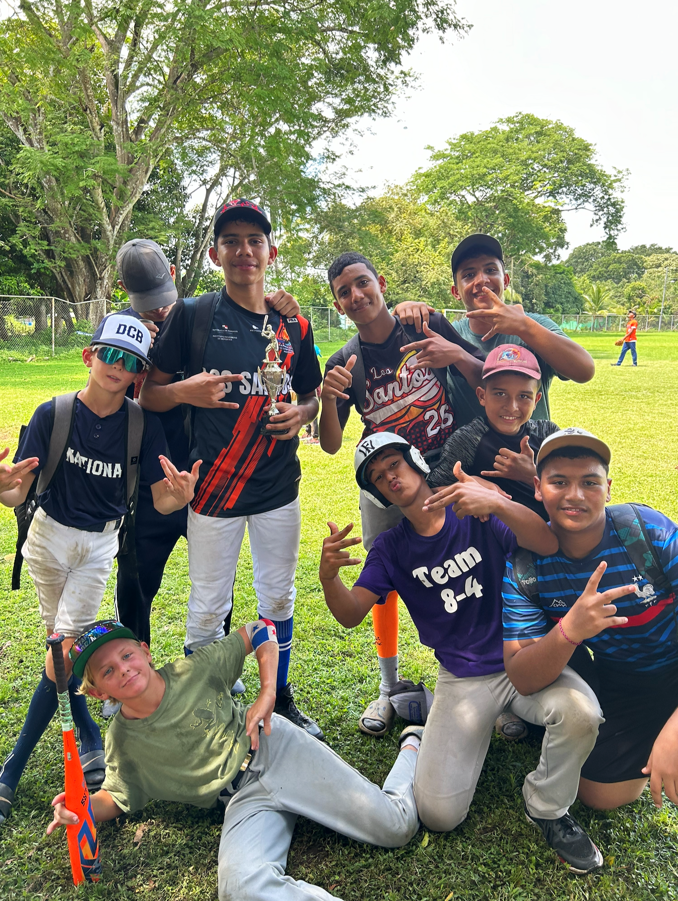 Group of young boys and teenagers at a park, some wearing sports gear, holding a trophy, posing and smiling for a photo on a grassy field with trees in the background.