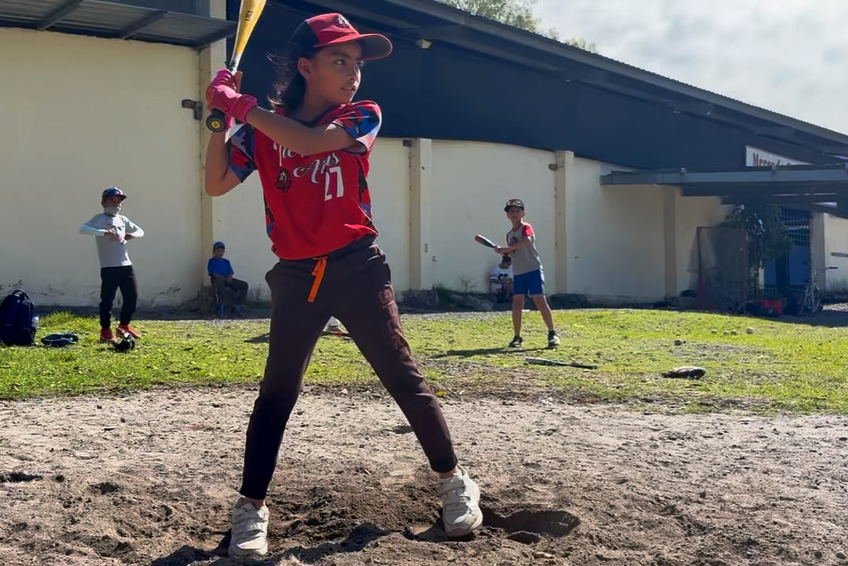 Young girl in red and black baseball uniform swinging a bat during practice, with other children in baseball gear in the background on a grassy and dirt field.