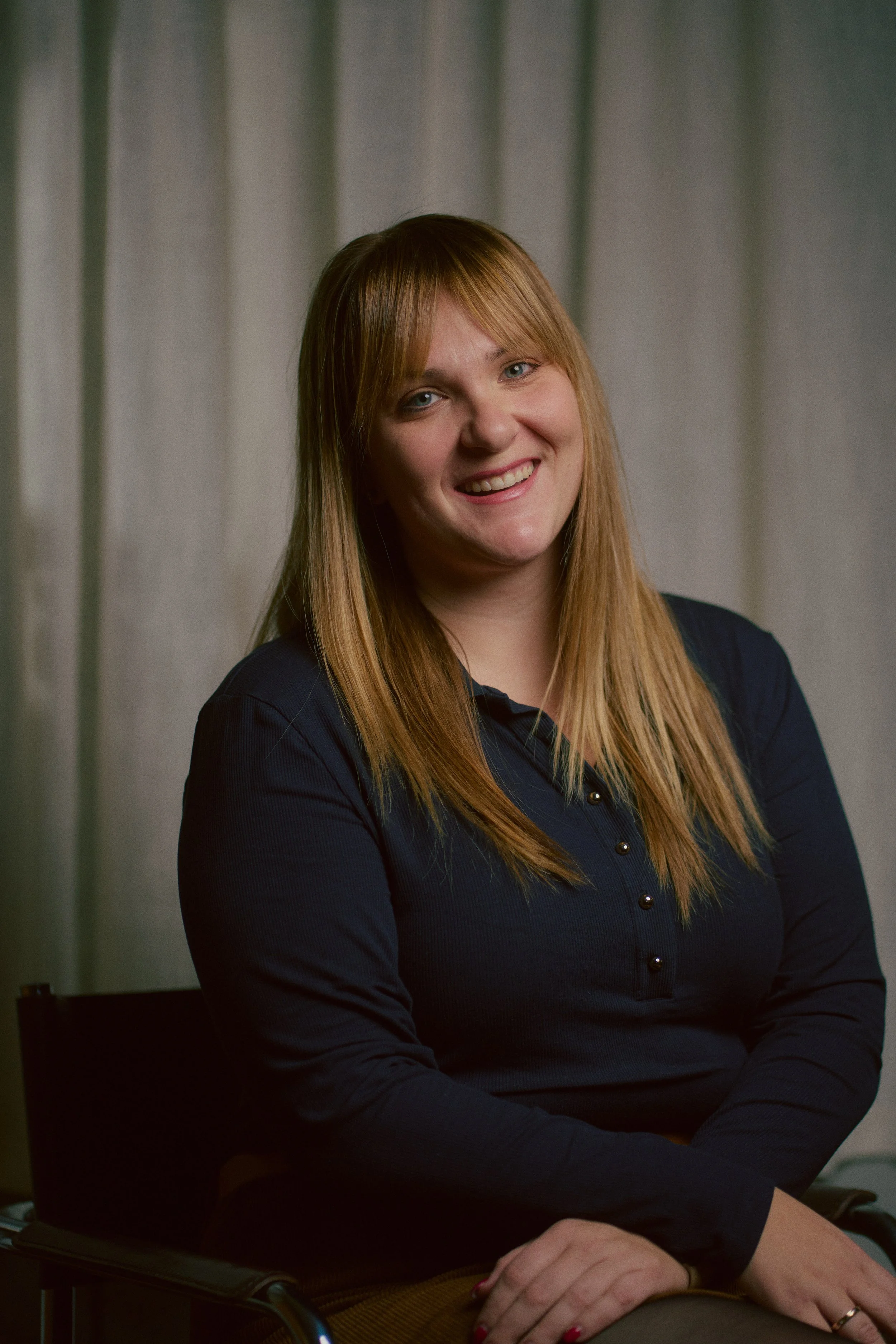 Ein Portrait einer lächelnden Frau mit langen, blonden Haaren, die ein dunkles Oberteil trägt, vor einem neutralen Hintergrund mit Vorhängen.