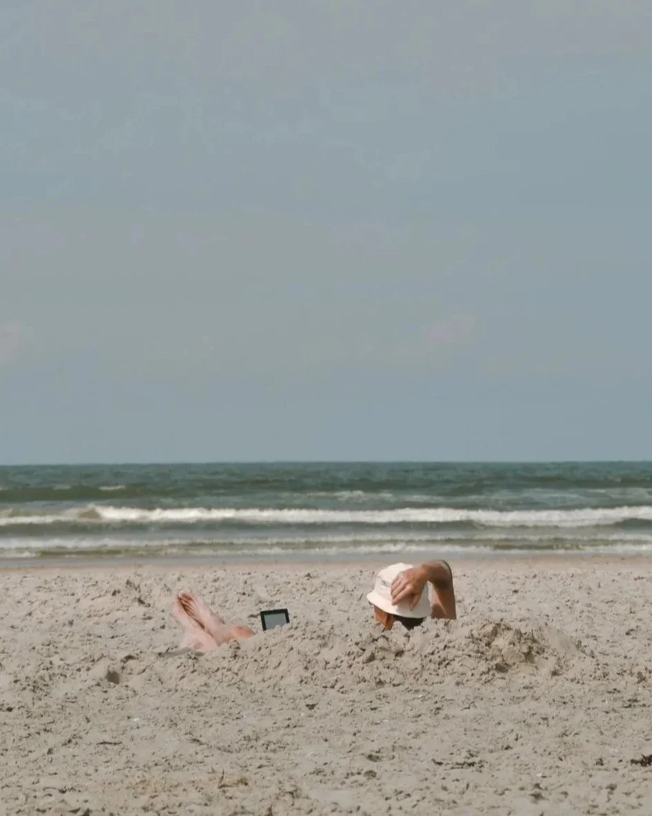Person liegt im Sand am Strand, trägt einen Sonnenhut und liest auf einem E-Reader, während die Füße aus dem Sand ragen. Im Hintergrund das Meer mit Wellen und ein leicht bewölkter Himmel.