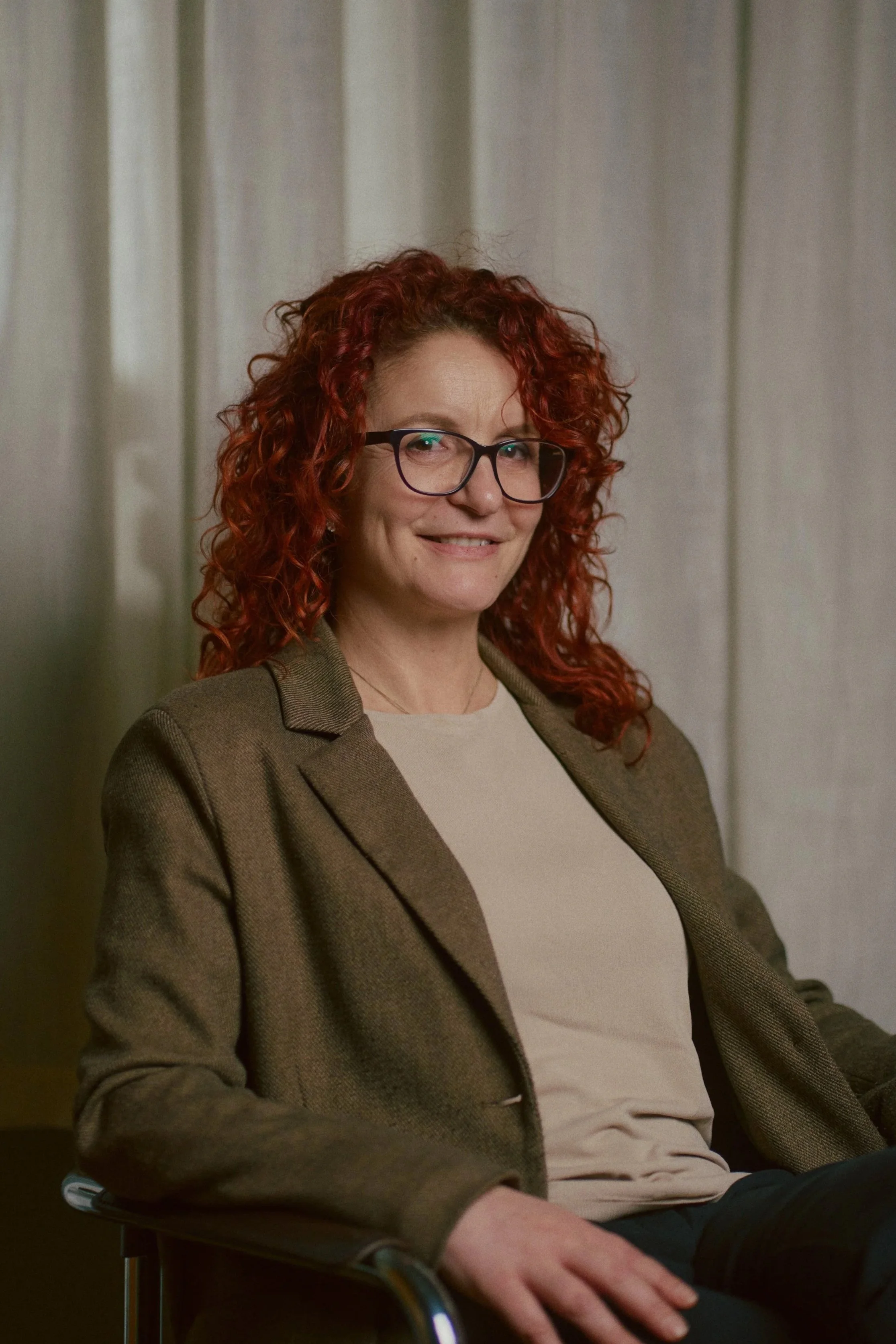 Eine Frau mit lockigem roten Haar trägt eine Brille und sitzt in einem Stuhl vor einem neutralen Hintergrund.