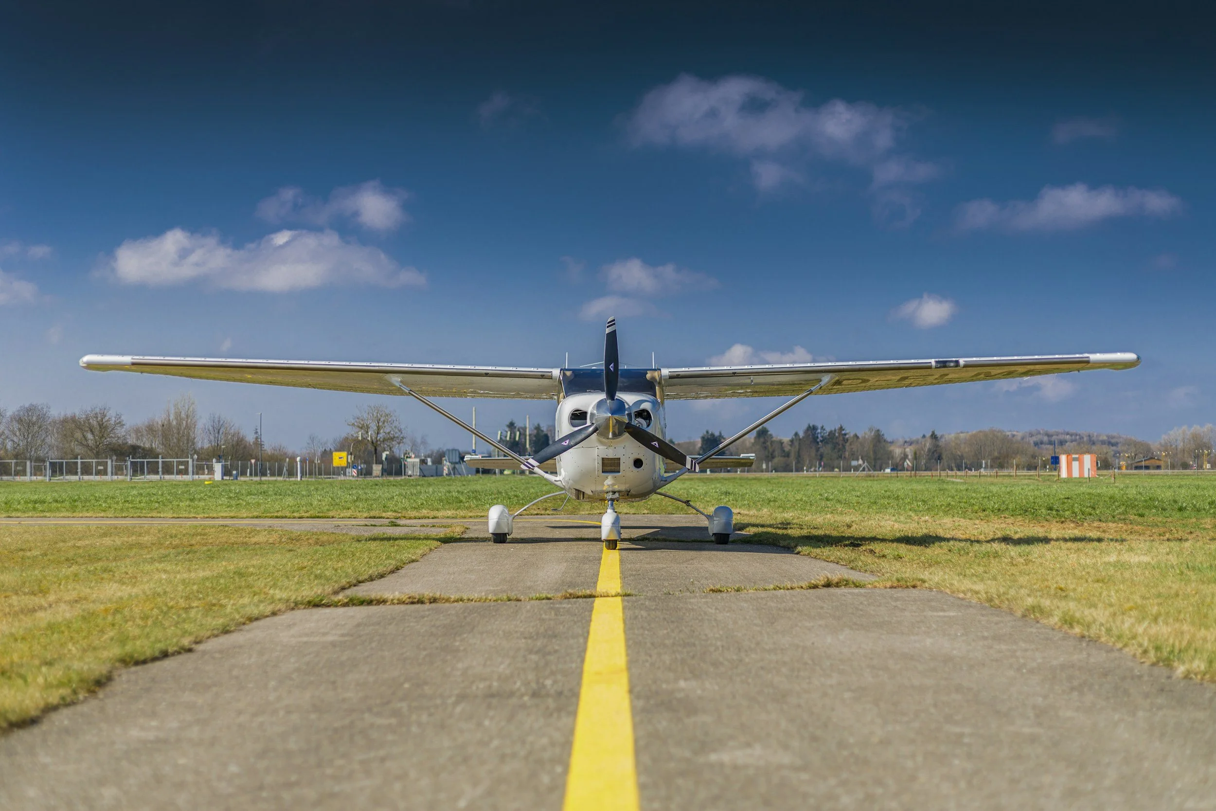 Small propeller airplane parked on runway with yellow line, grassy field, clear blue sky with scattered clouds, and distant trees.