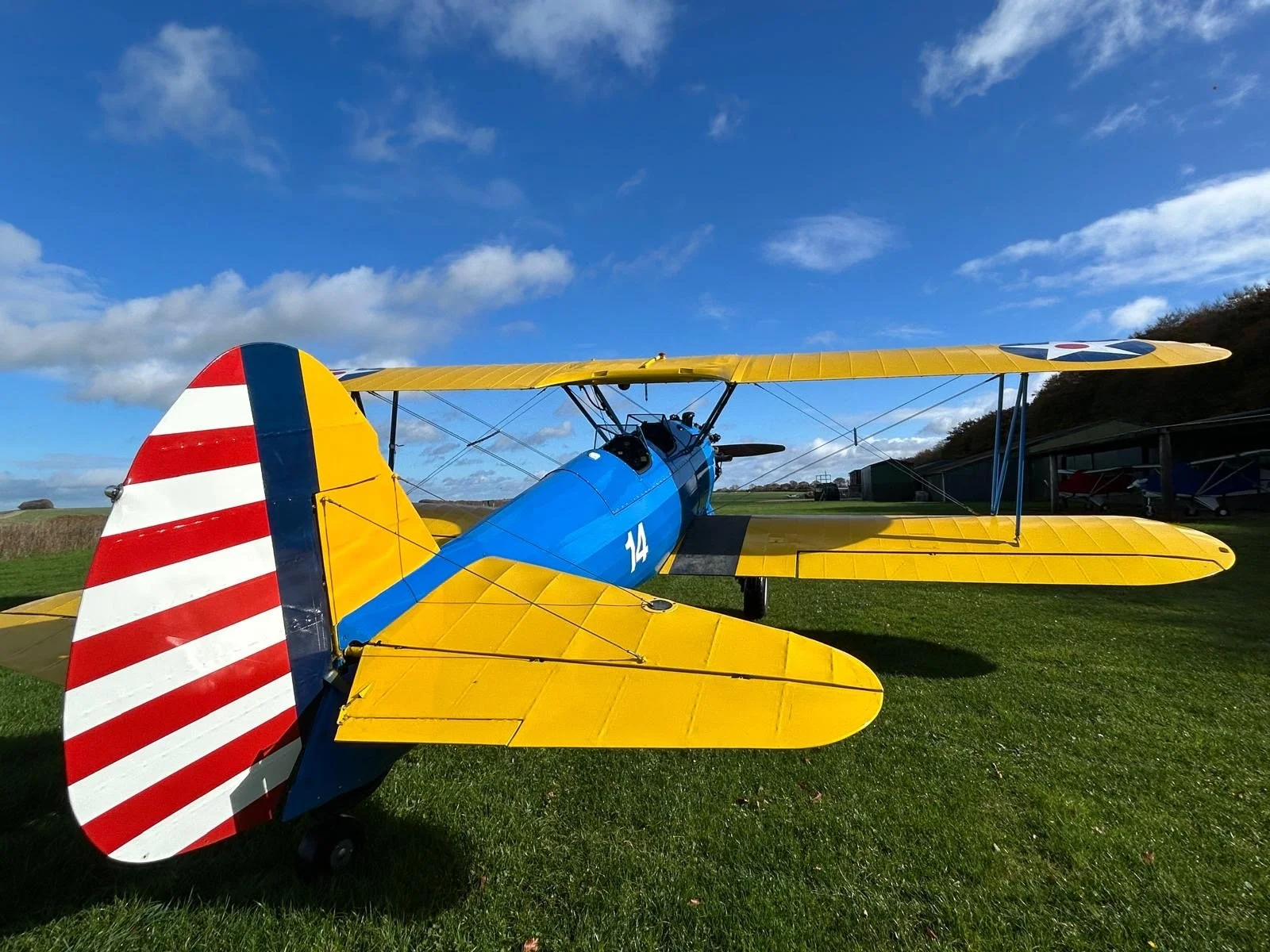 Small propeller airplane parked on runway with yellow line, grassy field, clear blue sky with scattered clouds, and distant trees.