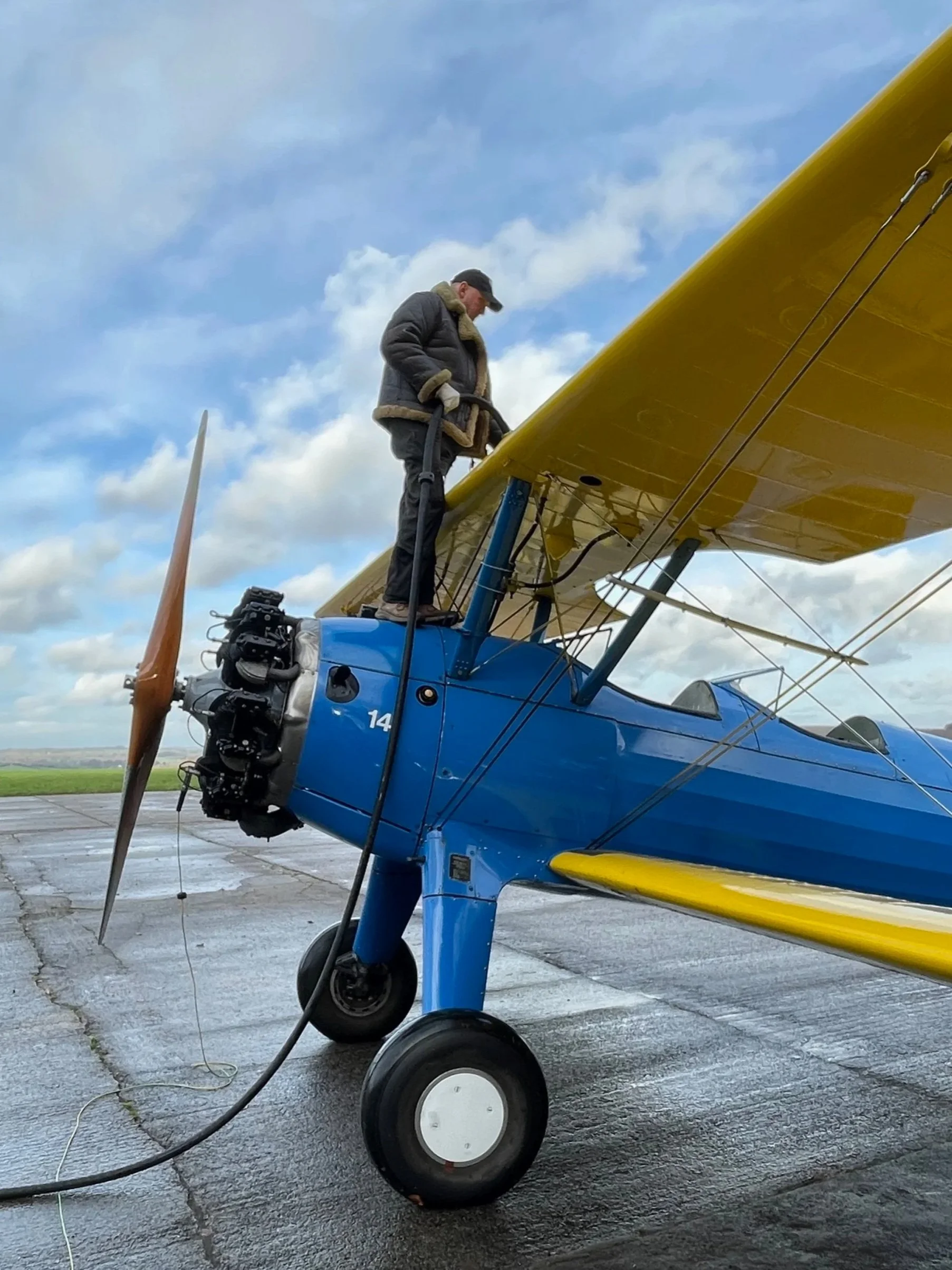 A person standing on the wing of a vintage blue and yellow biplane with a propeller, outdoors on a cloudy day.