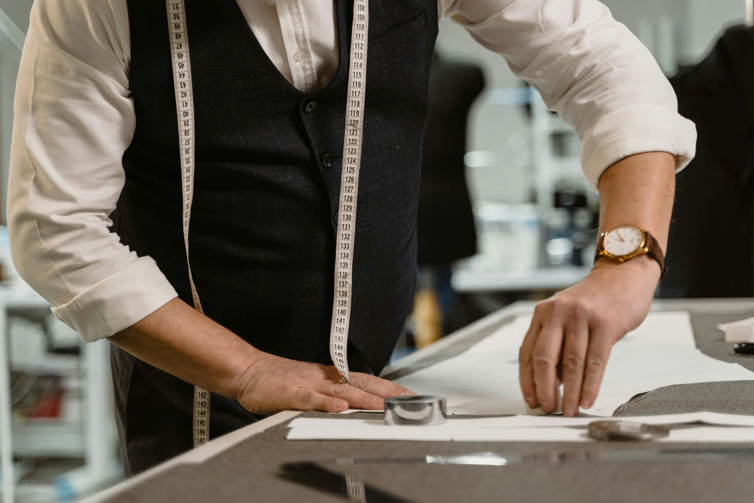 Person wearing a white shirt and dark vest measuring fabric with a measuring tape in a workspace.