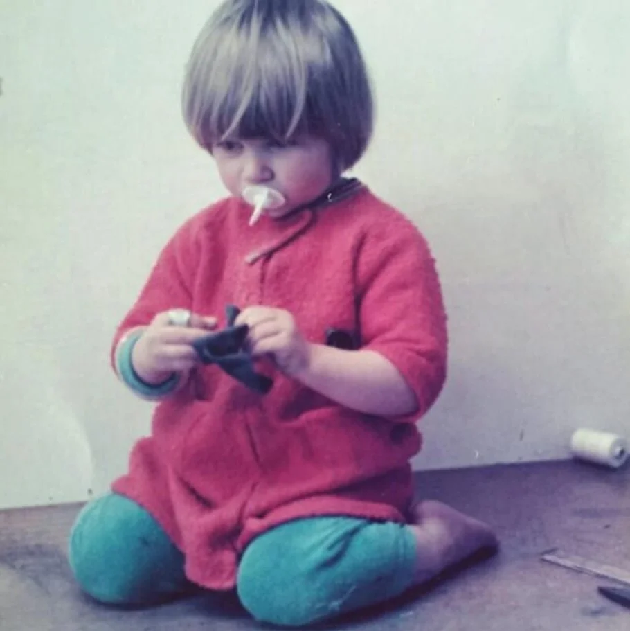 A young child with light brown hair kneeling on the floor, wearing a red jacket and teal pants, holding a small object, with a pacifier in their mouth.
