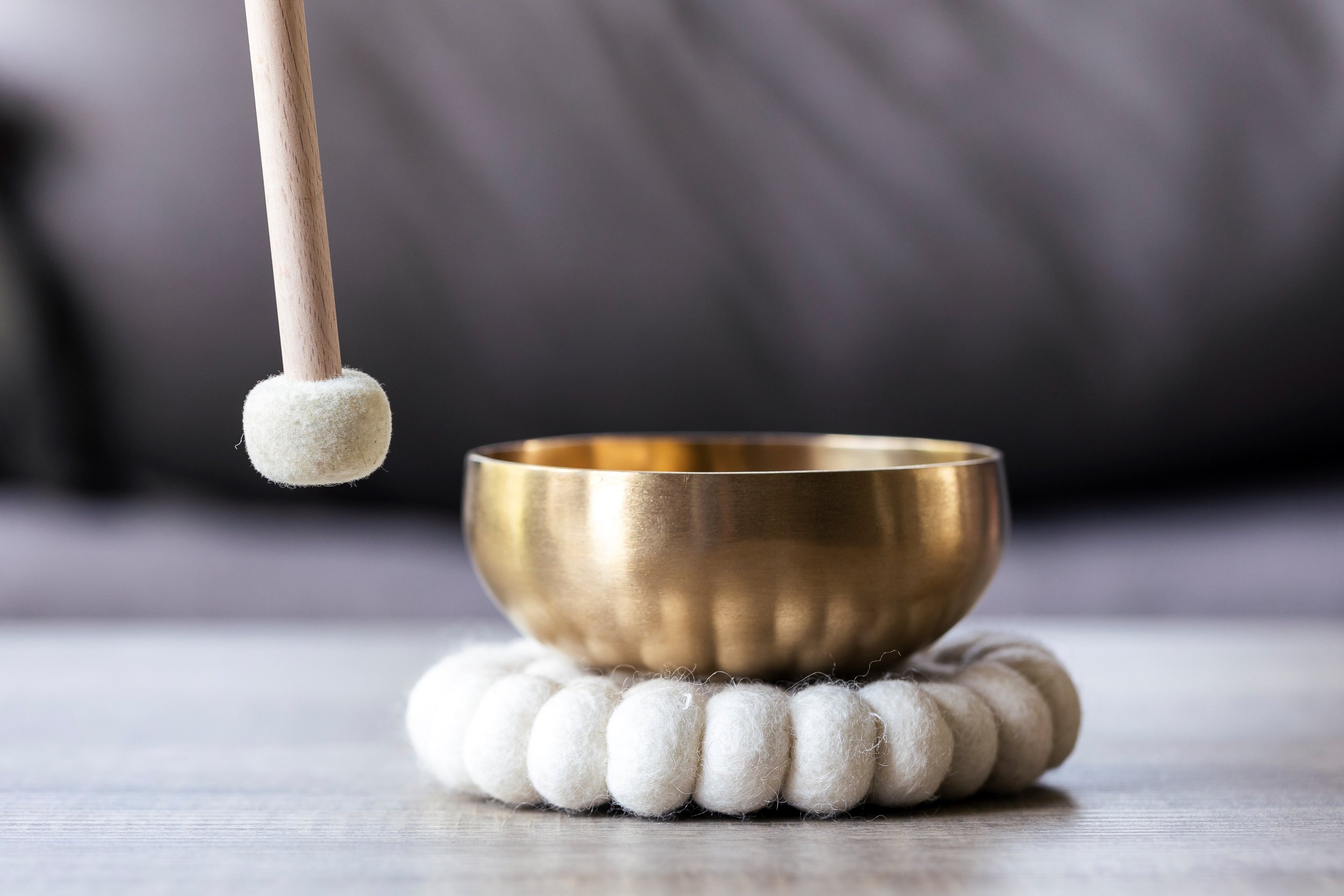 A brass singing bowl resting on a white felt circle with a wooden mallet suspended above it.
