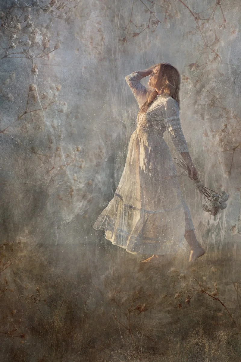 A woman in a vintage dress standing in a misty, foggy outdoor setting, holding a small bouquet of flowers, with her hand on her forehead, gazing into the distance.