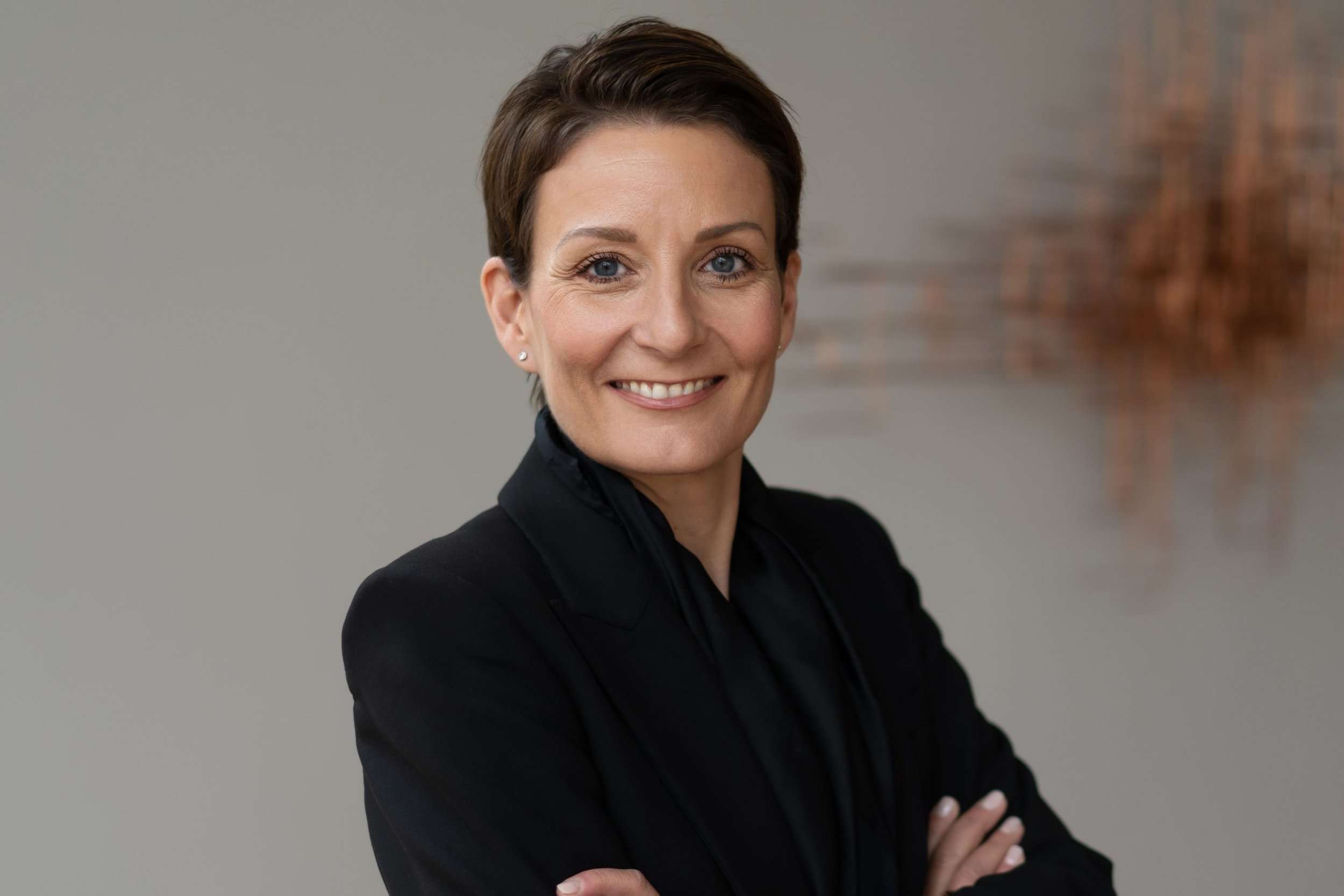 Die Gründerin von creda partners Patricia Schauß mit kurzen braunen Haaren, dunkler Kleidung und einem freundlichen Lächeln, vor einem grauen Hintergrund und kupferfarbenen Wanddekoration.
