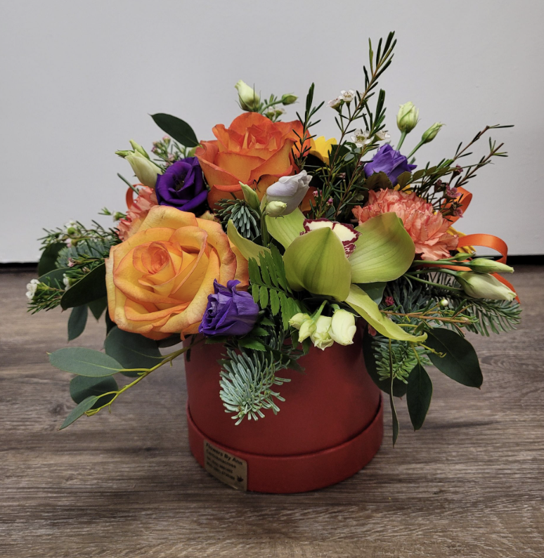 A colorful flower arrangement in a red round box, featuring orange roses, purple lisianthus, green orchids, carnations, various greenery, and small white flowers.