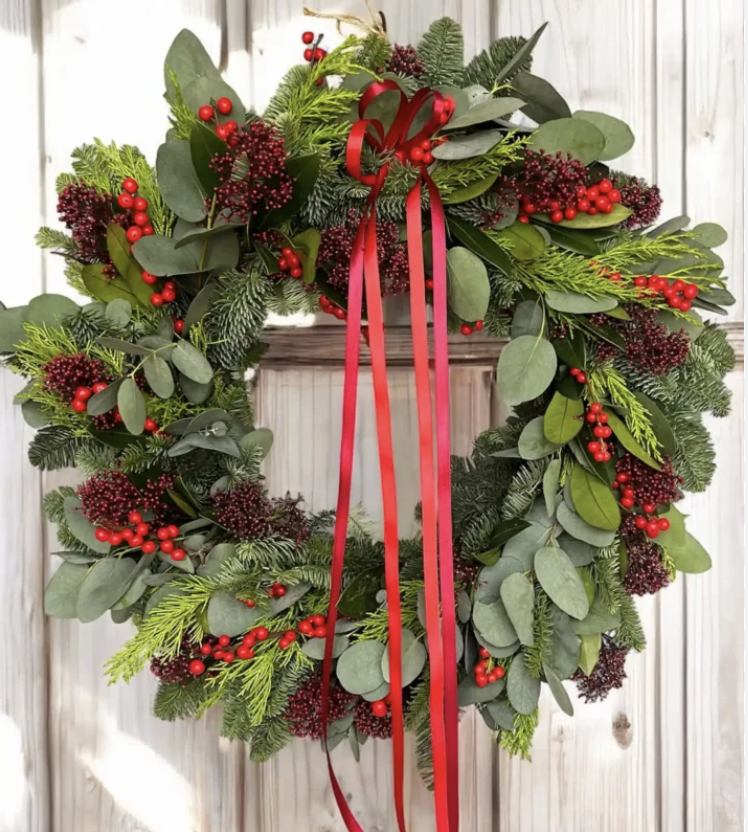 Decorative Christmas wreath with green foliage, red berries, and a red ribbon hanging in front of a white wooden background.