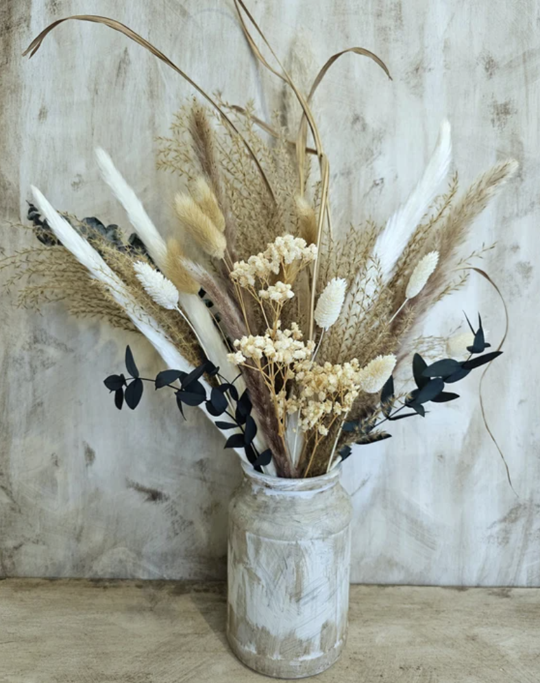 A dried flower arrangement in a rustic white ceramic vase, featuring pampas grass, dried bees, and eucalyptus leaves, set against a textured beige wall.