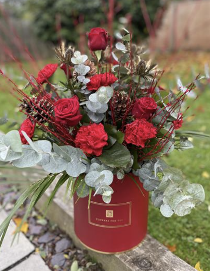 A red floral arrangement in a red box with gold accents, containing red roses, red carnations, white eucalyptus leaves, pinecones, and dried red twigs, outdoors on a wooden surface.