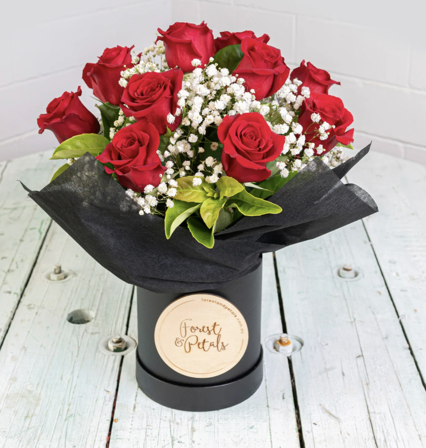 Bouquet of red roses with white baby's breath flowers and green leaves in a black wrapping and container, on a white wooden surface with a white brick wall background.