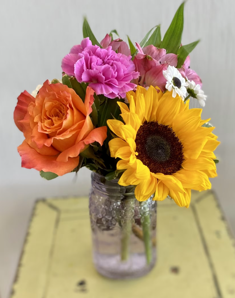 A glass jar with water holding a bouquet of colorful flowers including a large sunflower, pink carnations, an orange rose, and small white daisies, positioned on a yellow table.