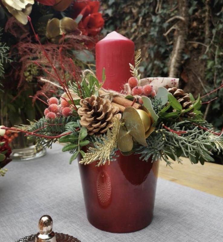 Christmas centerpiece with red candle, pinecones, red berries, gold pine branches, and greenery in a red vase.