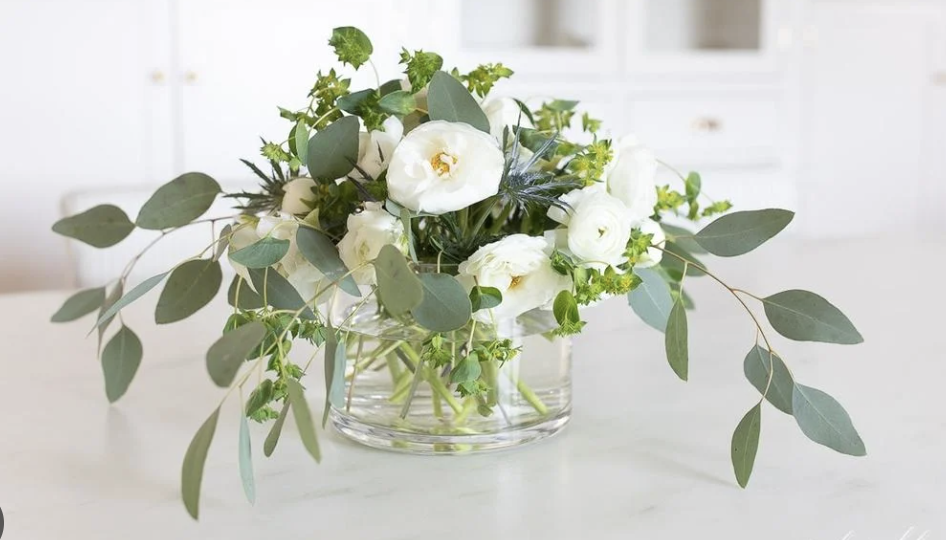 A clear glass vase filled with white flowers and green foliage on a white surface in a bright room.