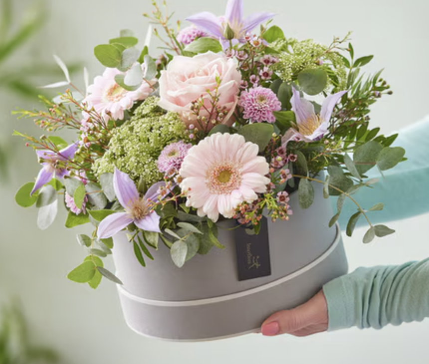 Hand holding a grey hatbox filled with pink and purple flowers, including roses, gerbera daisies, and lilies, with green foliage.