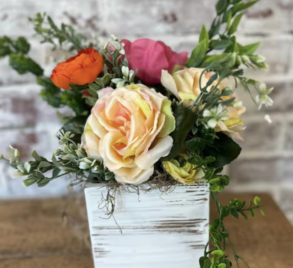 Colorful artificial flower arrangement in a white wooden vase on a wooden surface with a brick wall background.