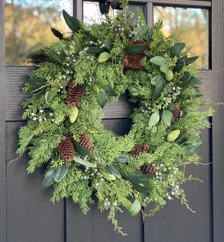 Fresh green holiday wreath with pinecones, greenery, and small white berries hanging on a black door.
