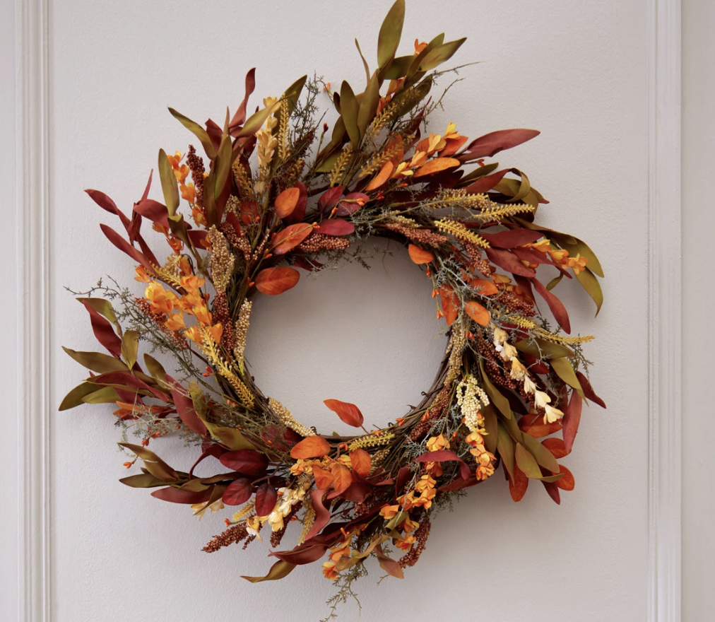 Fall-themed wreath with orange, red, and yellow leaves and flowers on a white wall.