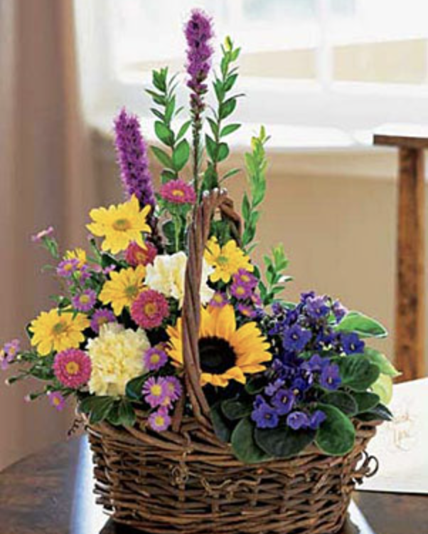 Colorful flower basket with sunflowers, purple, yellow, and pink flowers on a wooden table near a window.