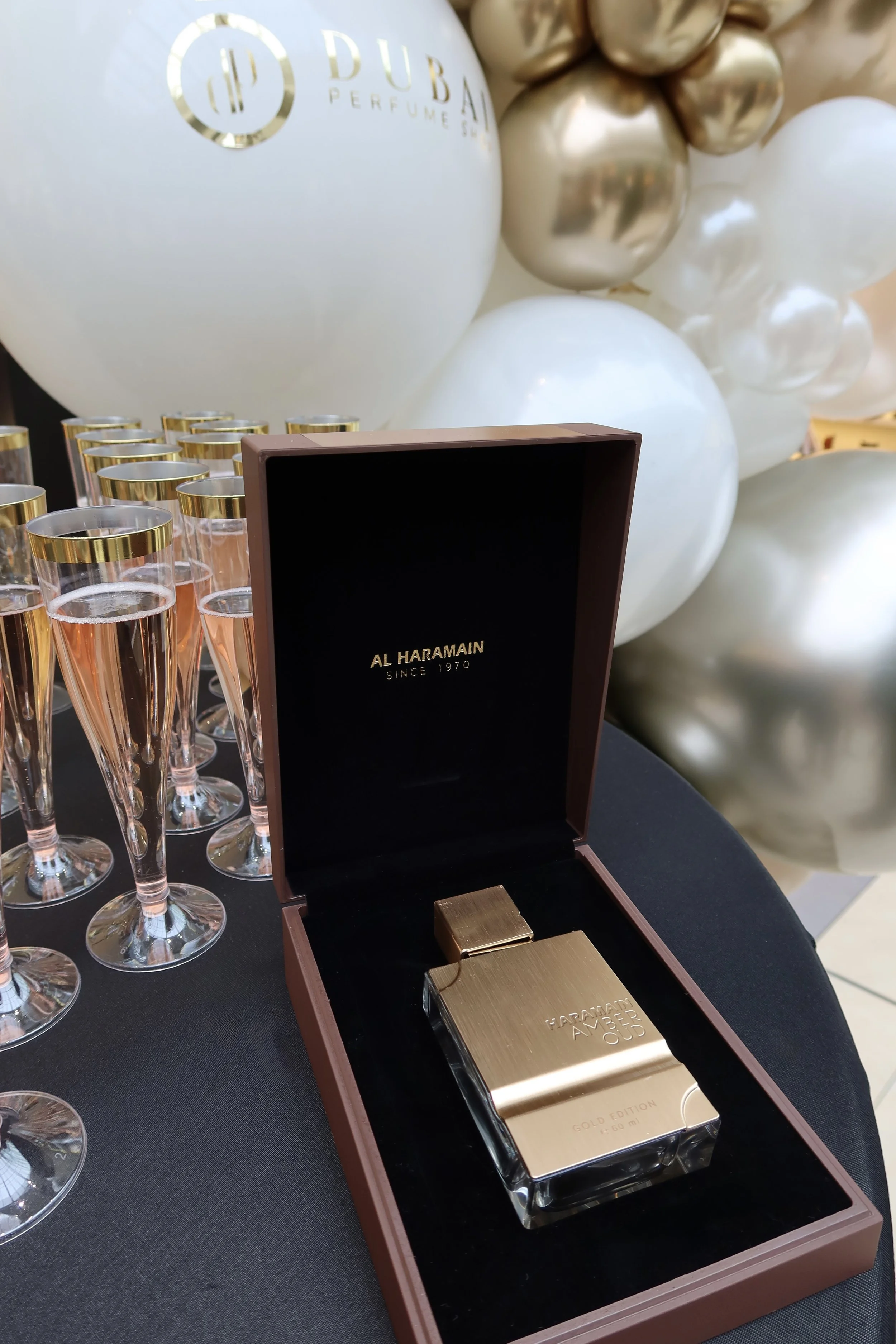 A perfume bottle inside a brown box with black interior, featuring gold lettering, surrounded by glasses filled with rosé champagne, in front of a balloon display at a Dubai Perfume Show.