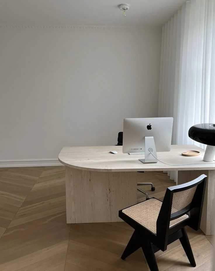 A minimalistic office with a light wood desk holding an Apple iMac, wireless mouse, and a touchpad. A black and beige chair is positioned in front of the desk, with sheer white curtains on the right side and a plain white wall in the background.