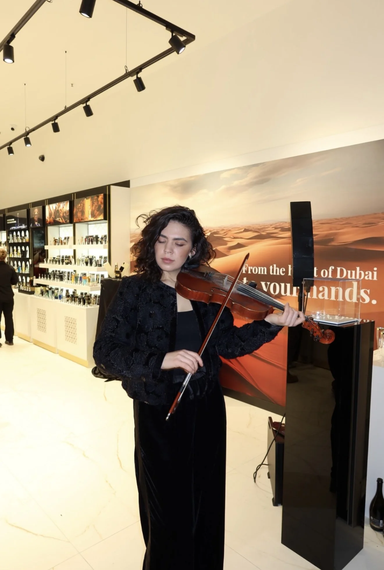 Woman playing violin indoors with a desert landscape backdrop and a display of bottles and items in the background.