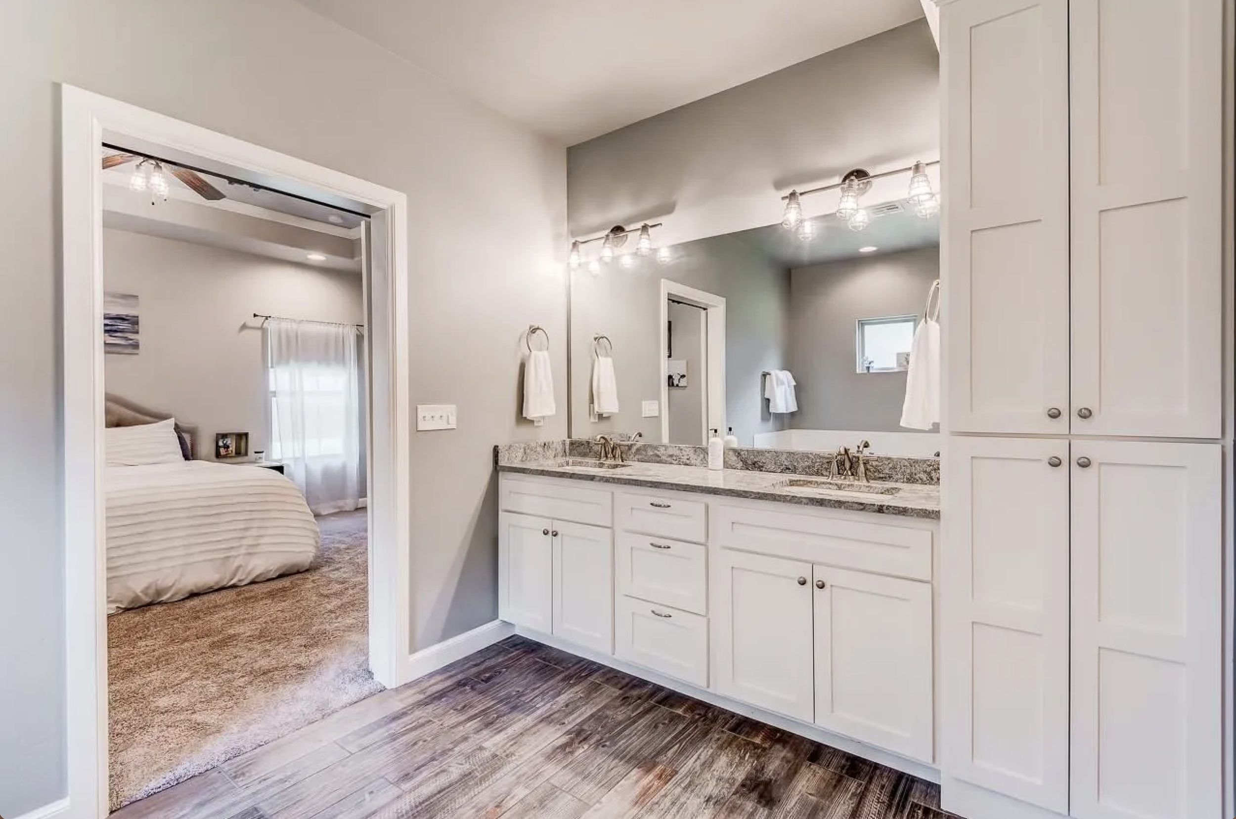 Bathroom with double vanity sink, granite countertops, large mirror, overhead lighting, white cabinetry, and separate toilet and shower area visible in the background. Bedroom with a bed and window with curtains visible through doorframe.