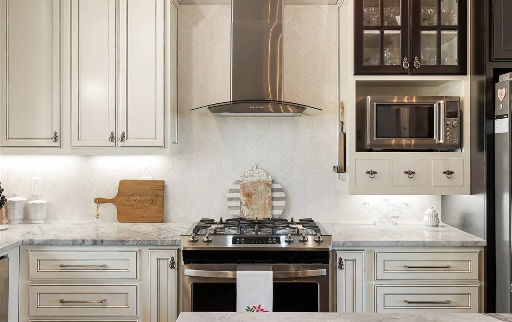 Kitchen with white cabinetry, marble countertops, stainless steel stove and oven, microwave, and modern range hood. Decor includes a small cutting board, a white lamp, and a decorative plaque.
