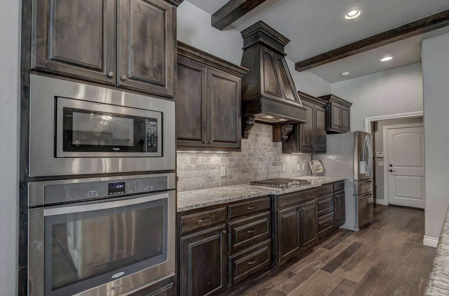 Modern kitchen with dark wood cabinets, stainless steel microwave and oven, granite countertops, brick tile backsplash, and wooden flooring.
