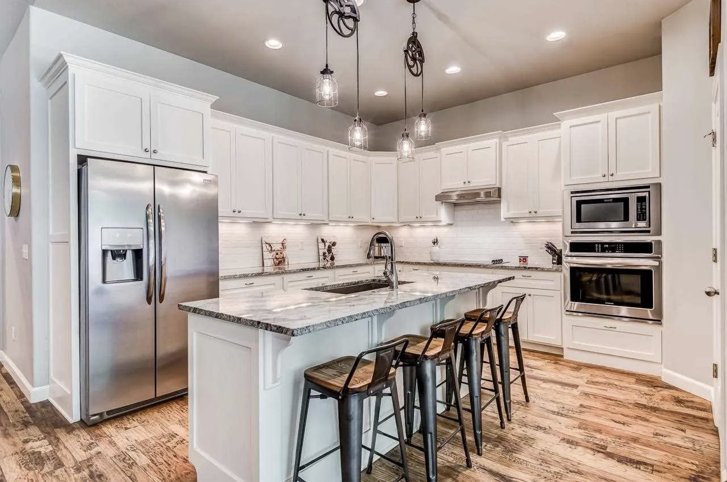 Modern kitchen with white cabinets, granite countertops, stainless steel refrigerator, double oven, microwave, and hanging lights over an island with four stools.
