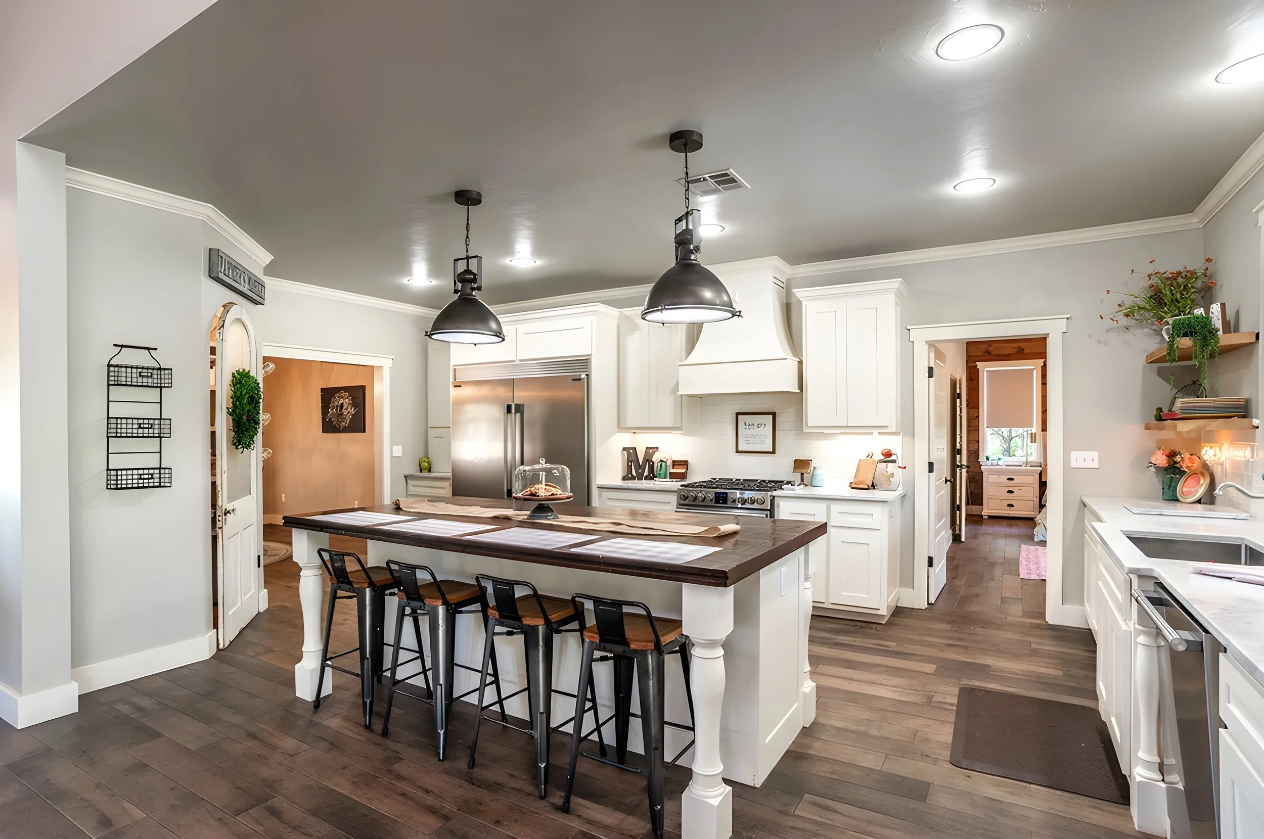 Modern kitchen with white cabinetry, a large island with a dark wood top, four black stools, stainless steel refrigerator, and hanging black pendant lights.