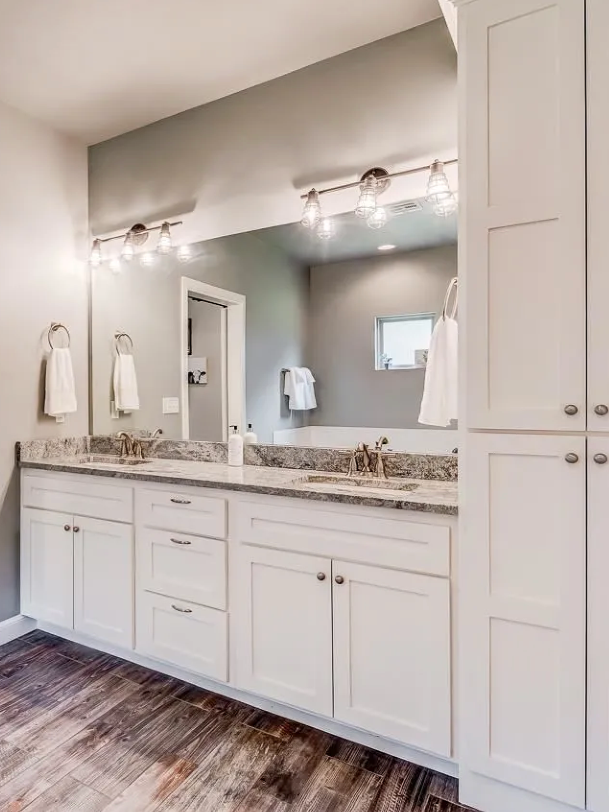 Modern bathroom with white vanity, granite countertop, and dual sinks, illuminated by overhead lighting and natural light from a small window.