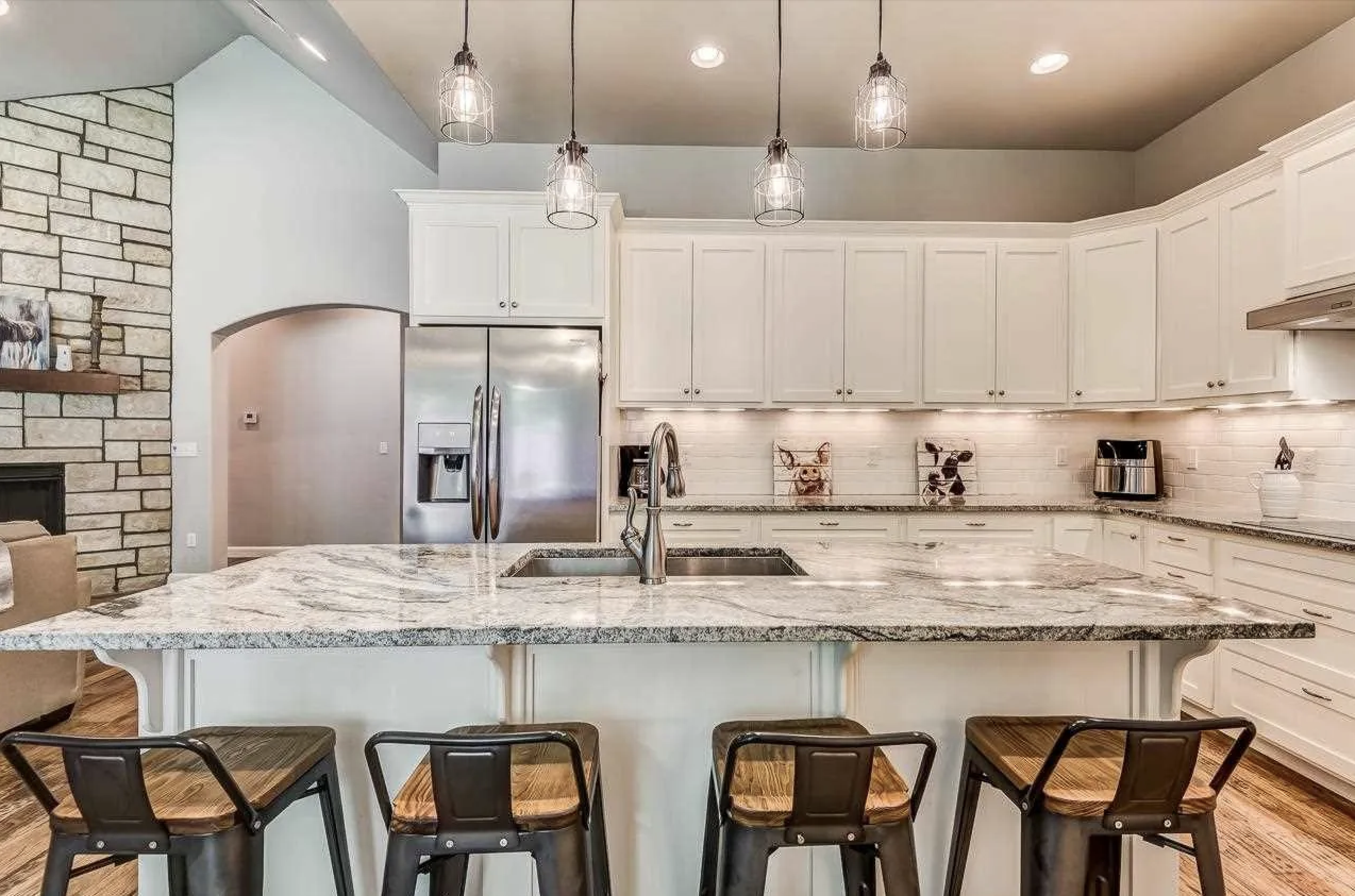Modern kitchen with white cabinets, granite countertops, stainless steel refrigerator, island with a sink, three bar stools, granite backsplash, pendant lighting, and decorative artwork.