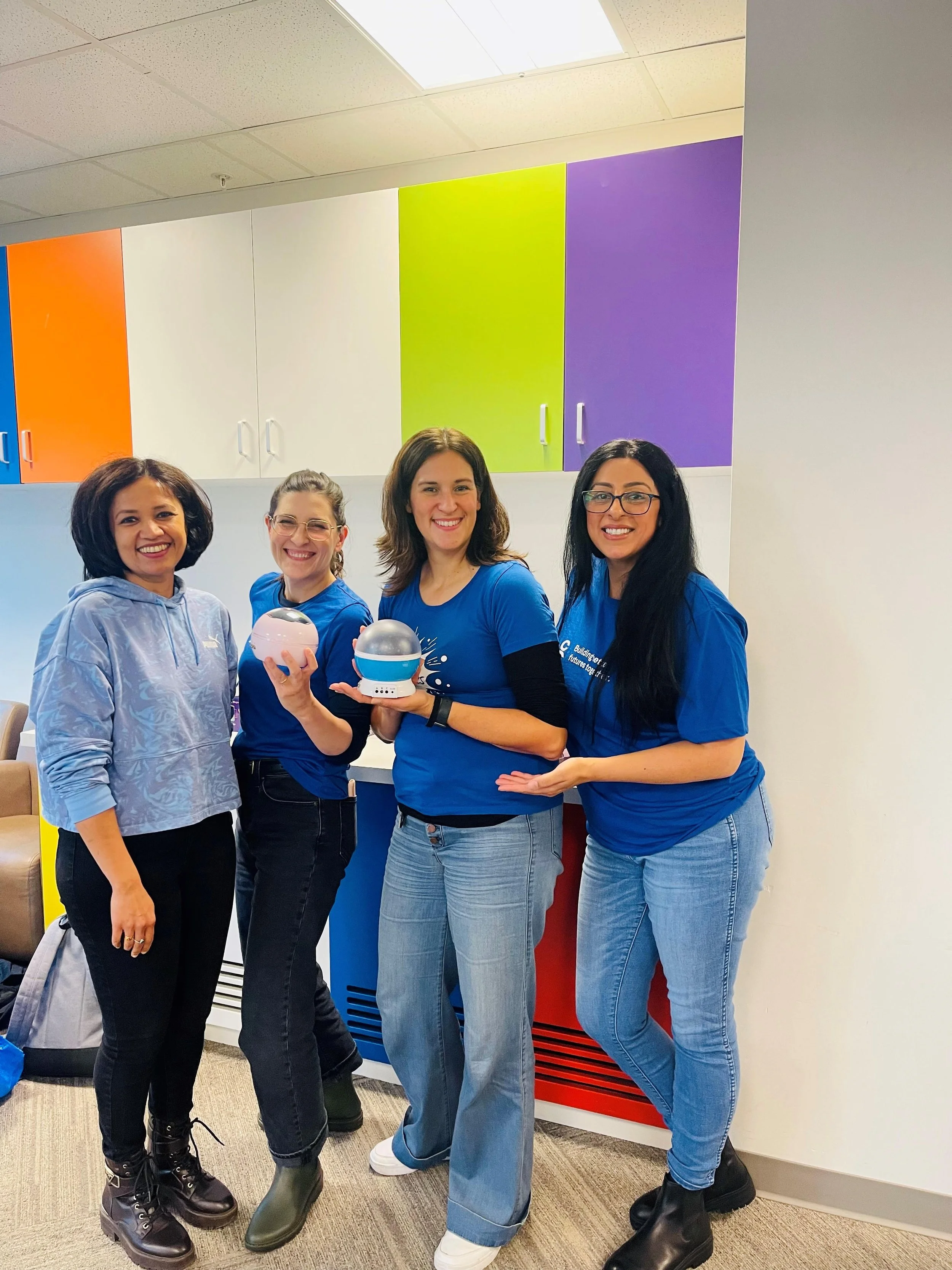 Four women standing in front of colorful lockers, smiling, with two women holding spherical objects resembling robots or devices.