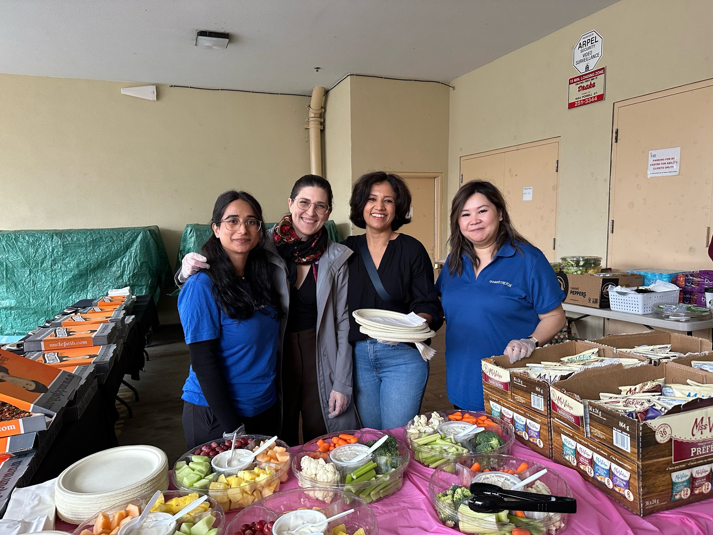 Four women standing behind a table with trays of cut vegetables and snacks at a food drive or community event.