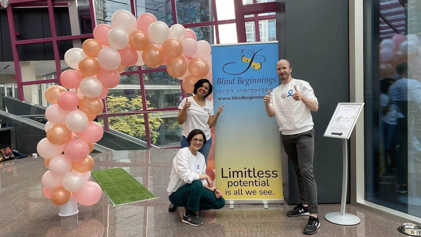 Three people standing and squatting in front of a blue and yellow banner for Blind Beginnings, with a balloon arch made of pink and peach balloons to the left. Two women and one man are smiling, giving thumbs up or a thumbs down. The background shows
