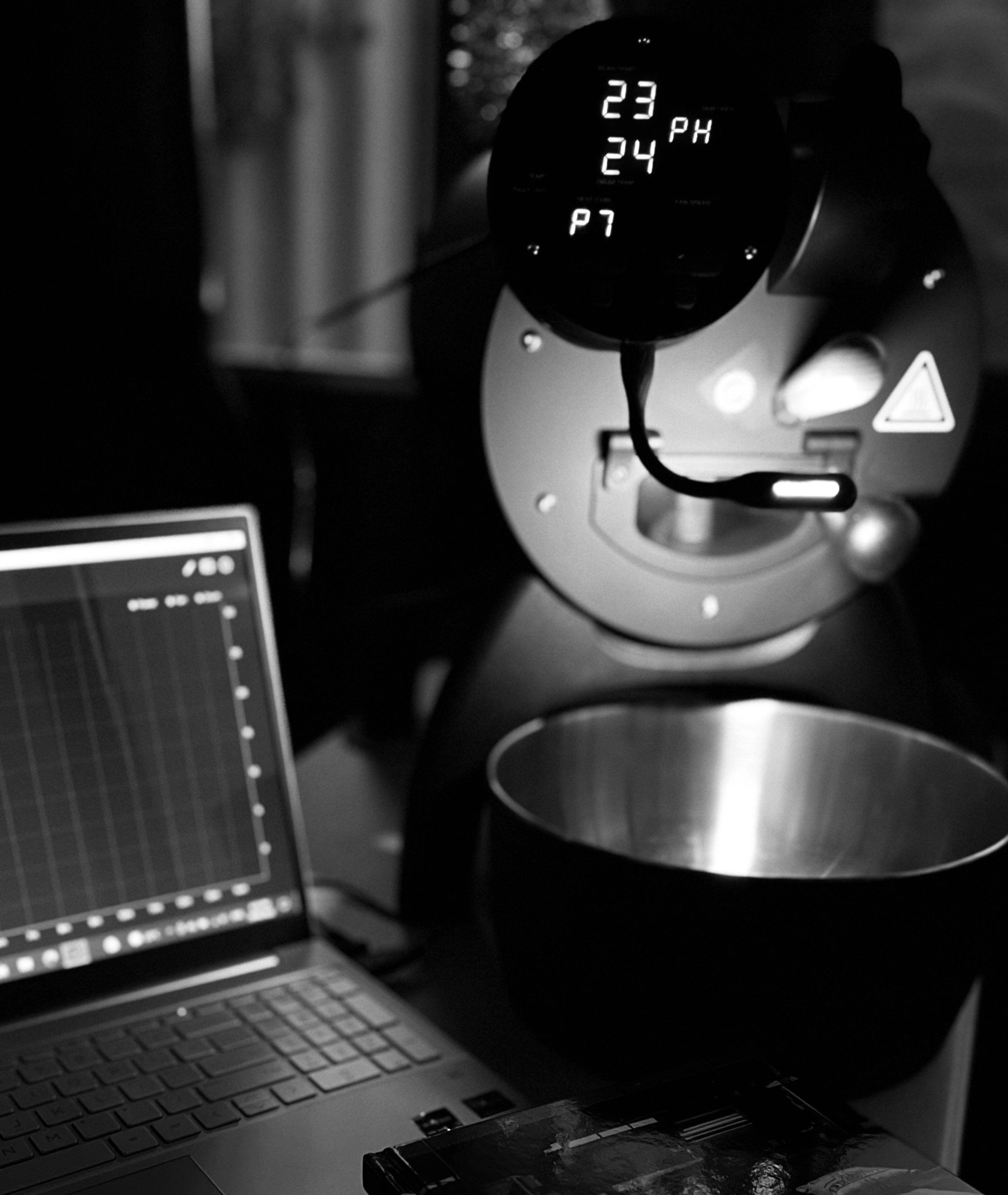 A black and white photo of a coffee roaster with a digital display showing temperature, a laptop with a graph, and a metal cooling tray underneath the roaster.
