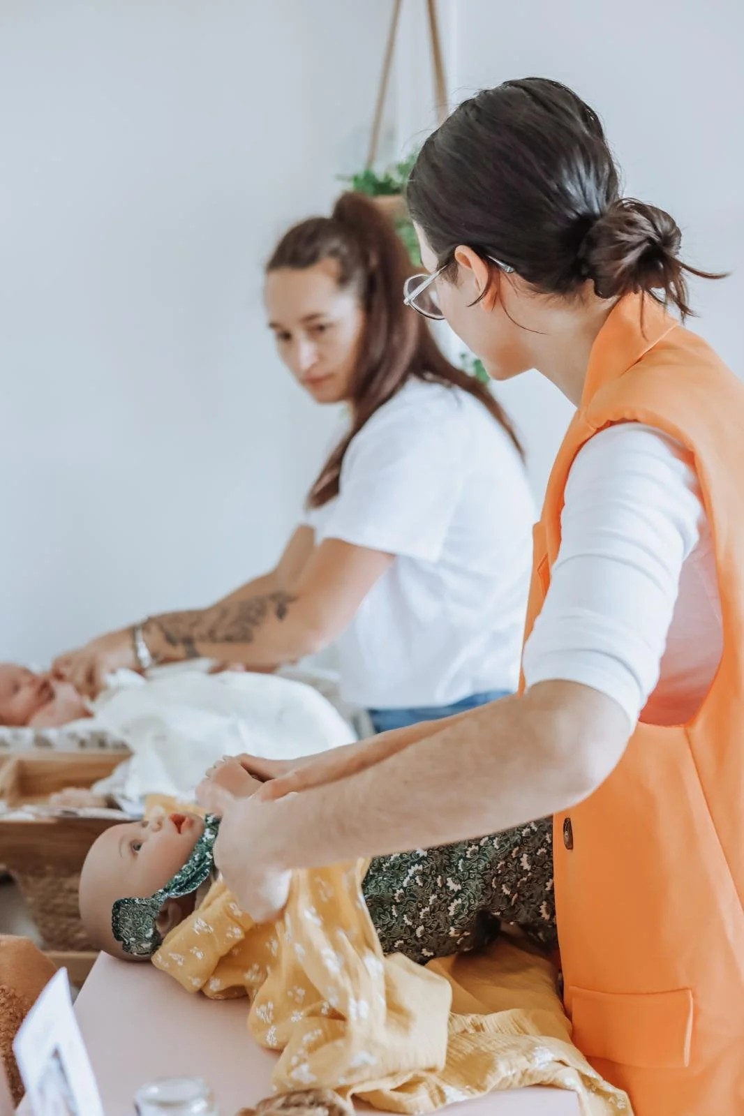 Deux femmes et un bébé dans un environnement apparemment de soins ou d'activités artisanales, avec un décor intérieur simple et des objets en bois et textile.