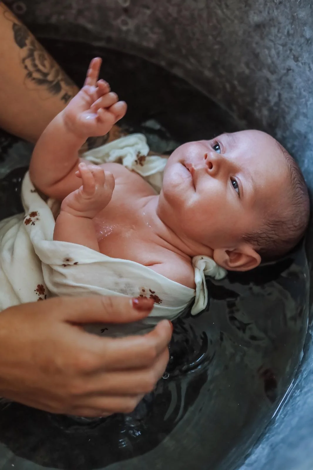 Un bébé en bain dans de l'eau noire, tenu par une personne dont les mains sont visibles, portant une chemise noire et ayant un tatouage sur le bras, regardant vers la caméra.