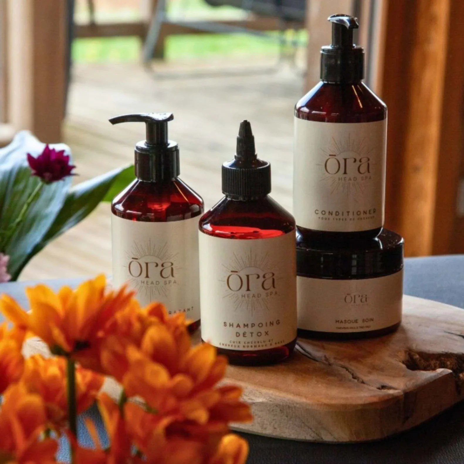 Bouteilles de produits capillaires de la marque Ôра, comprenant un shampooing detox, un masque et un conditionneur, placées sur une table en bois avec des fleurs orange en premier plan.