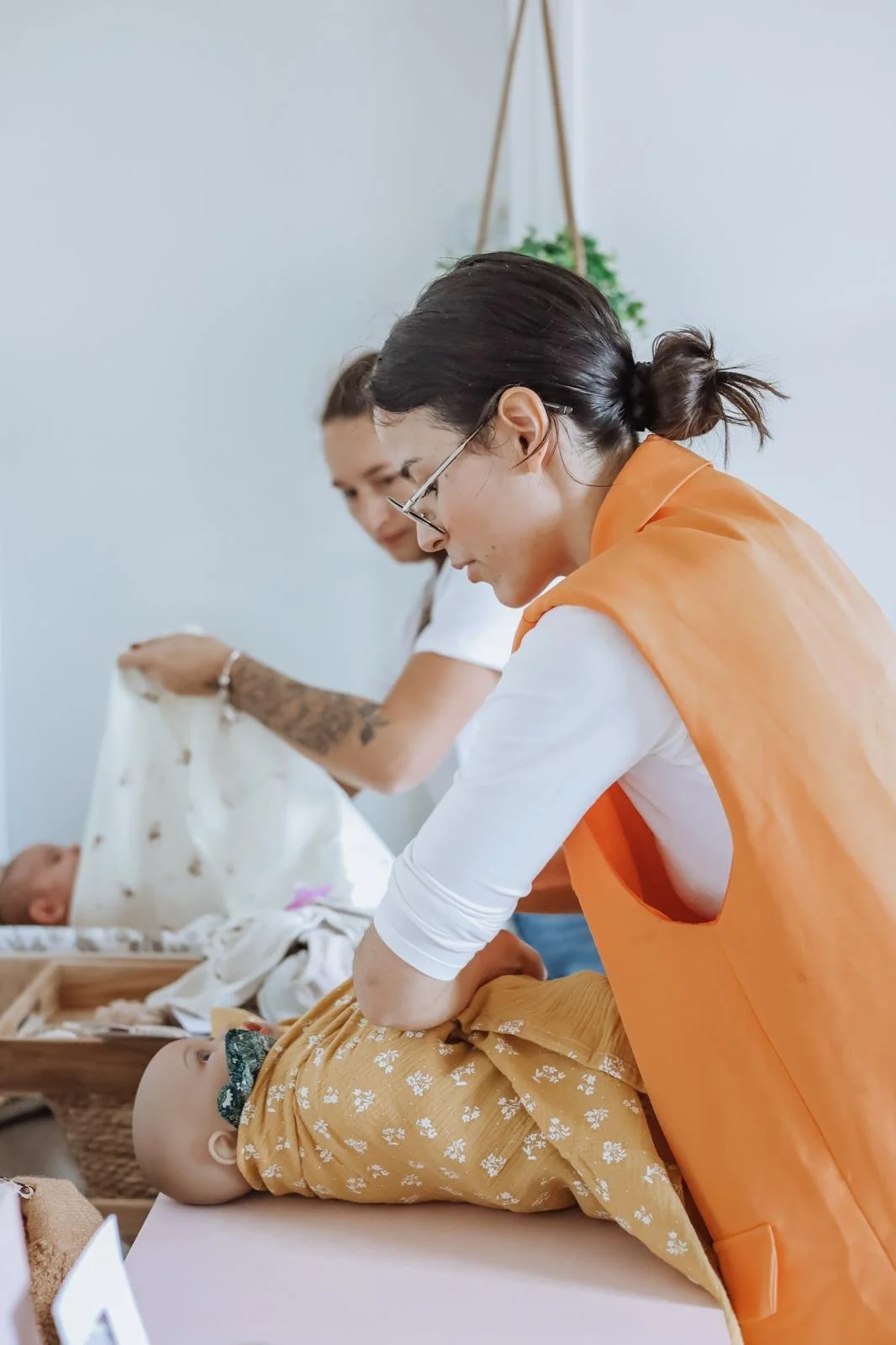 Une femme donne un bain à un bébé allongé sur une table, pendant qu'une autre femme la regarde en arrière-plan, dans un environnement intérieur calme.