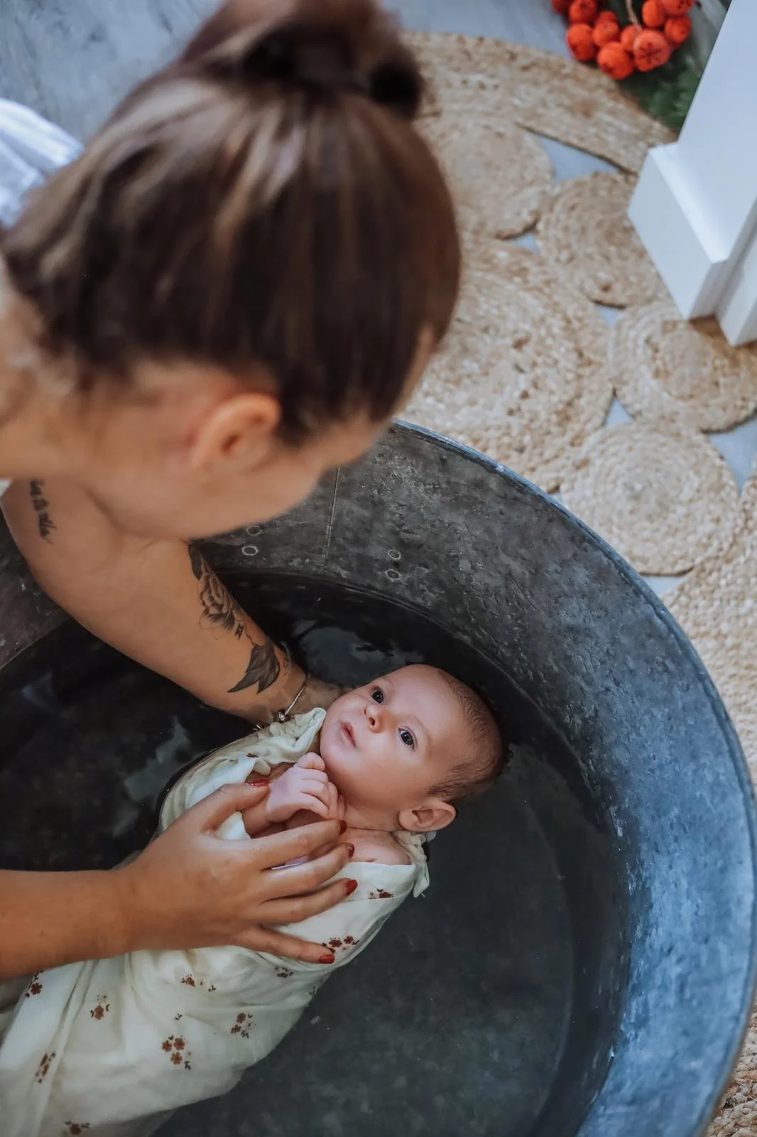 Une femme tient un bébé dans une baignoire, en train de lui donner le bain. Le bébé regarde vers la caméra, enveloppé dans une serviette beige. Il y a un tapis en forme de fleurs dans la pièce.