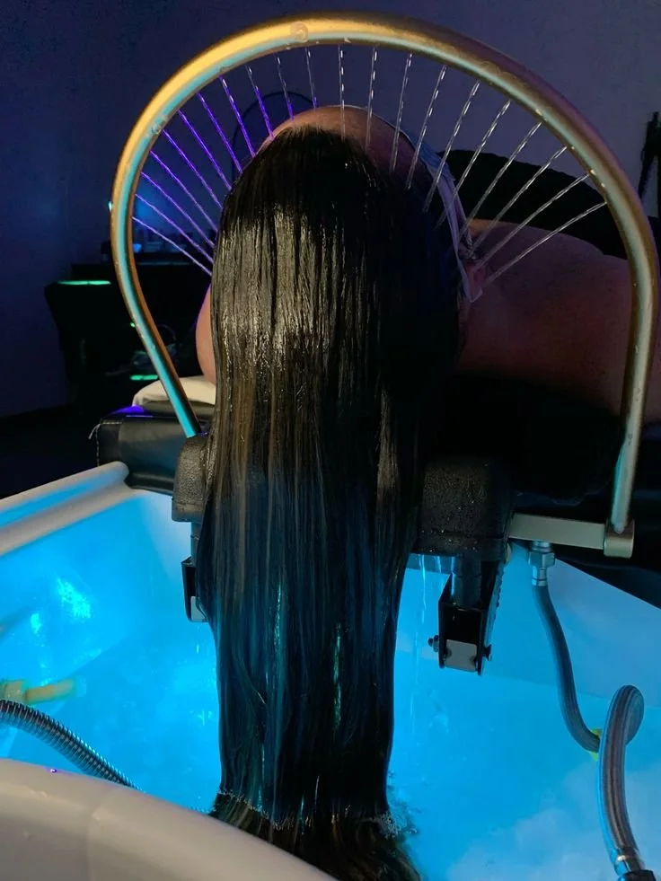 Une personne est sous une cabine de soins capillaires avec des cheveux longs et noirs, éclairée par des lumières colorées, dans une pièce sombre.