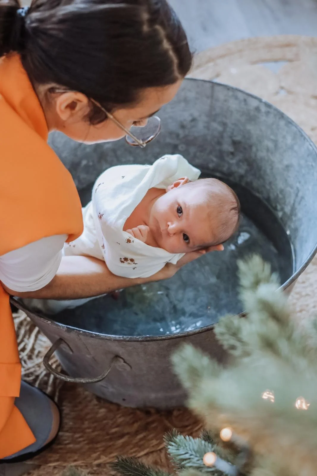 Un bébé dans un évier en acier inoxydable, étant tenu par une femme portant des lunettes et une chemise orange, dans un lieu intérieur.